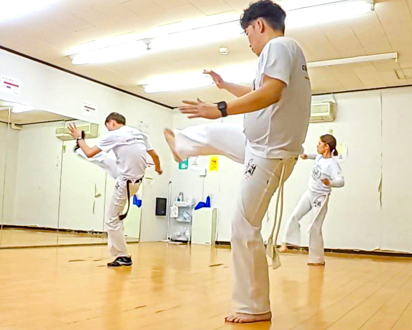 教室の会場で、白い道着を着た三人が鏡の前に立ち、脚を上げる基本の動きを練習している様子